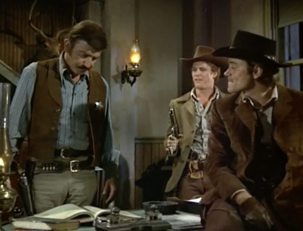 Three men in the gear of the Old West stand or sit around a desk in the sheriff's office.