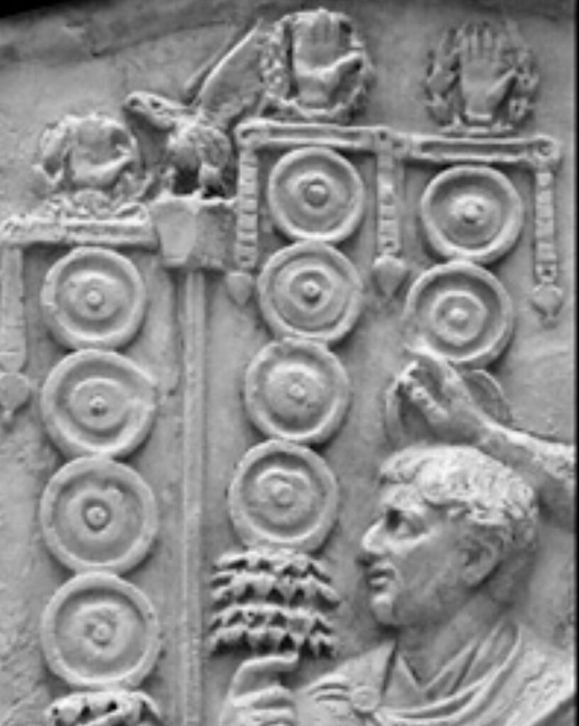 In the foreground, a Roman soldier’s head and shoulders; in the background, a row of four Roman military standards, three topped by the manus (hand symbol in a victory wreath), one by the aquila (legionary eagle).