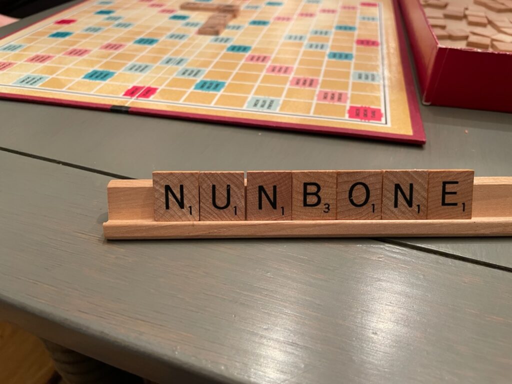 In the background, a Scrabble board. In the foreground, a Scrabble rack with 7 tiles showing: N, U, N, B, O, N, E.