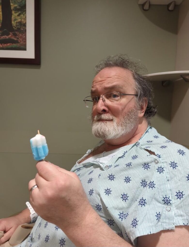 an old man whose hair has apparently never been combed sits in a hospital gown and holds aloft in weary triumph a half-eaten popsicle