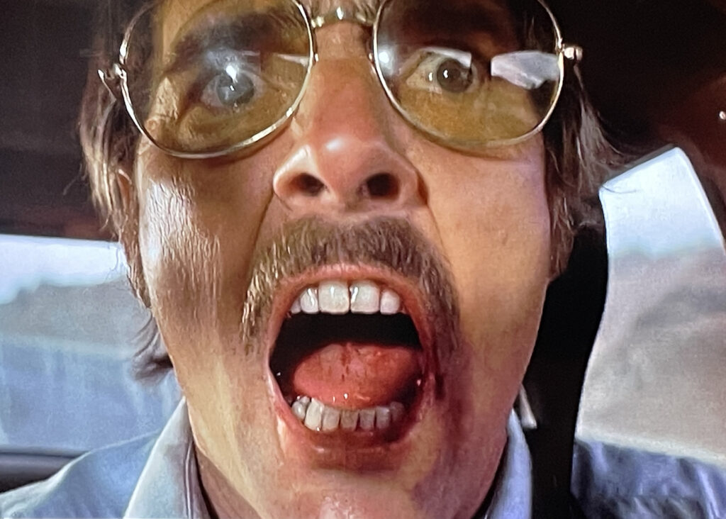 A still from DUEL; Dennis Weaver, wearing 70s-style aviator glasses gives the camera an open-mouthed scream.