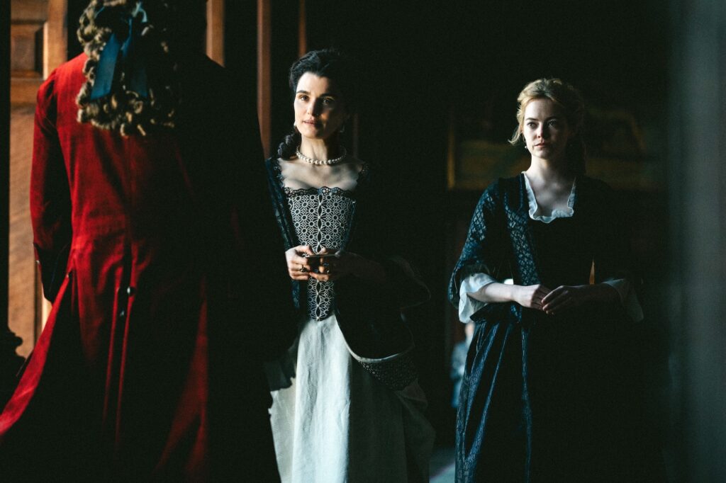 screenshot from Lanthimos' THE FAVOURITE (2018)
In the foreground: an official in a red coat with his back to us. Beyond him: Rachel Weisz as Lady Sarah Churchill (later the Duchess of Marlborough) and Rachel Stone as Abigail Hill (later Abigail Masham, still later Baroness Masham).